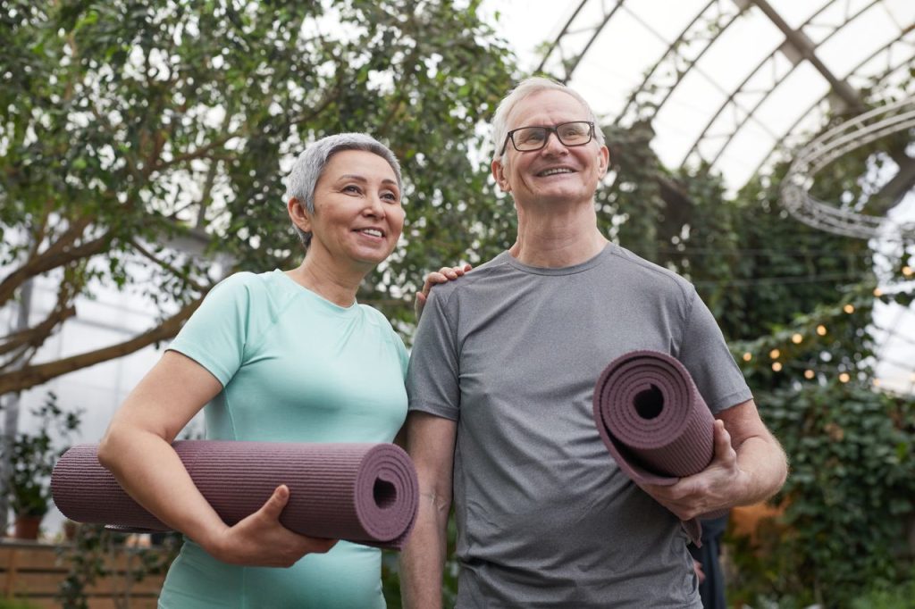 Happy senior couple holding yoga mats in a lush greenhouse setting.