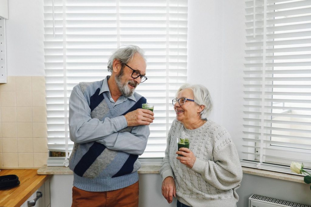 happy-senior-couple-standing-in-kitchen-with-smoothie-drinks-6975158 Senior couple smiling and enjoying green smoothies in a cozy kitchen setting.