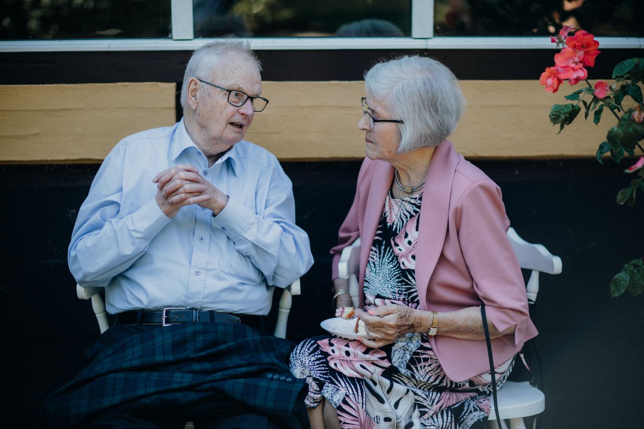 Un couple de personnes âgées assis à l'extérieur, en pleine conversation, entouré de fleurs. Une scène prise sur le vif.